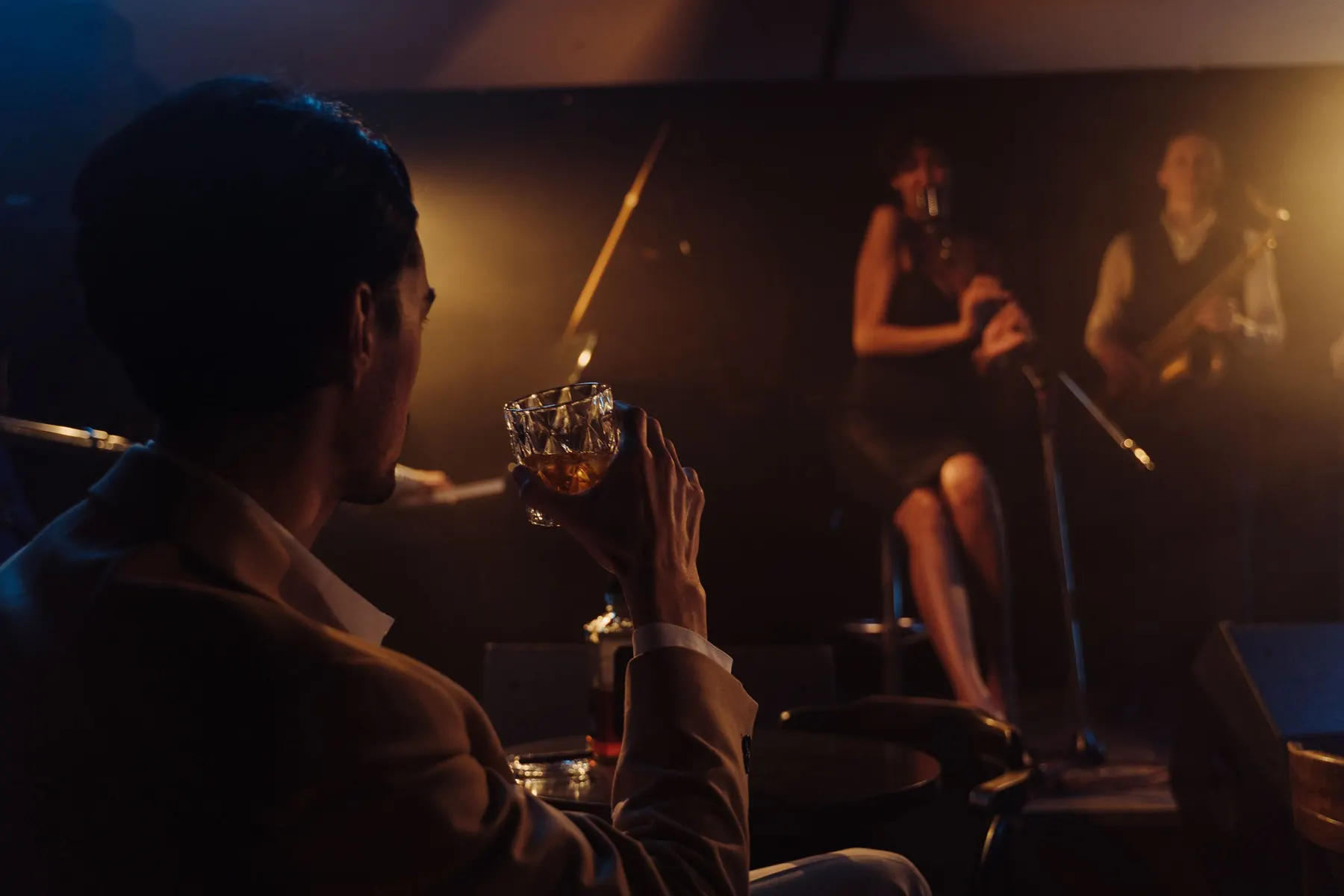 A man in a brown jacket holding a glass of whisky, seen from behind, watching a jazz band on a lit stage beyond a small table.