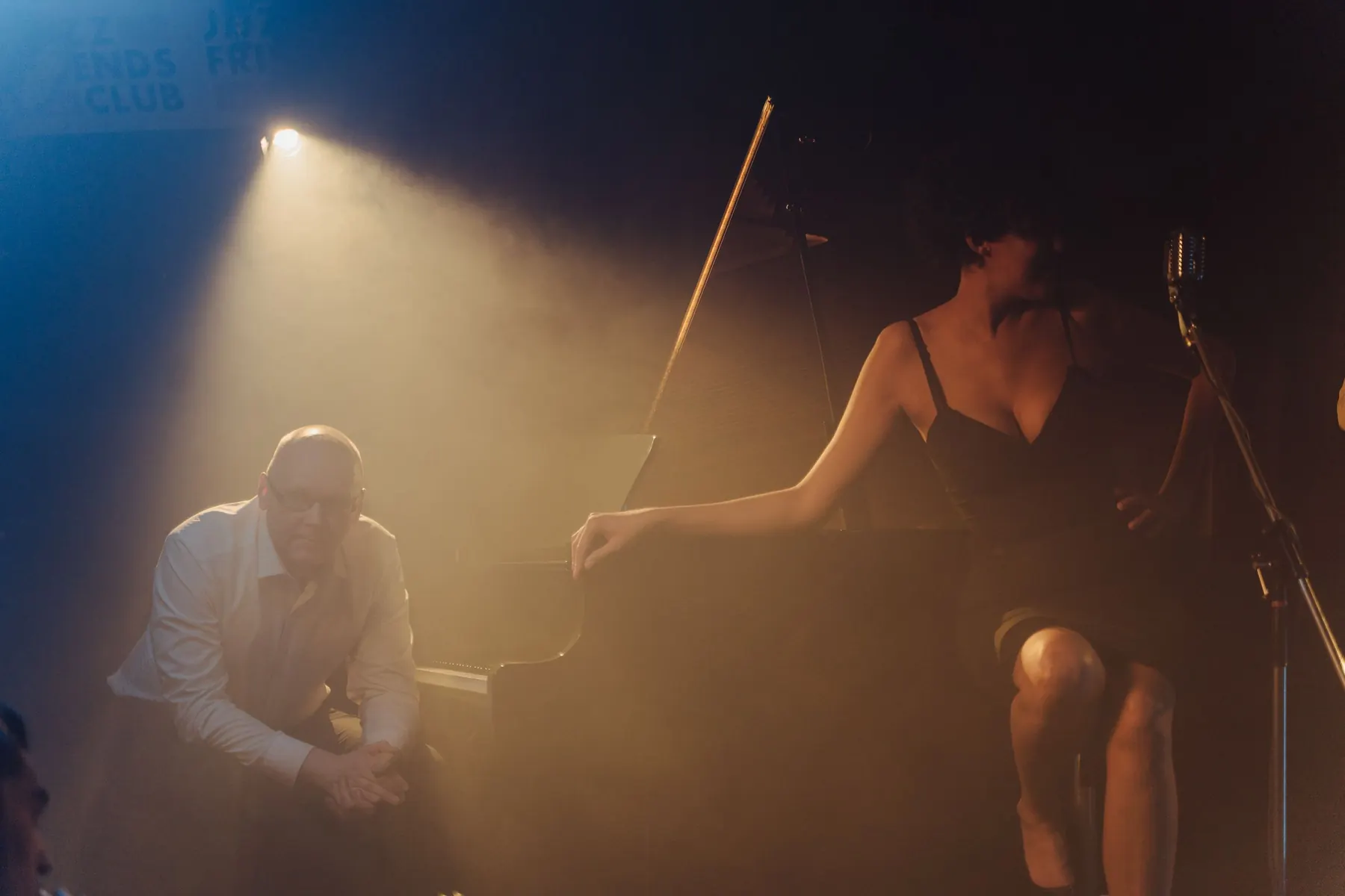A pianist leaning toward the camera beside a grand piano while a singer in a black dress sits on the piano lid, lit by a shaft of yellow stage light.